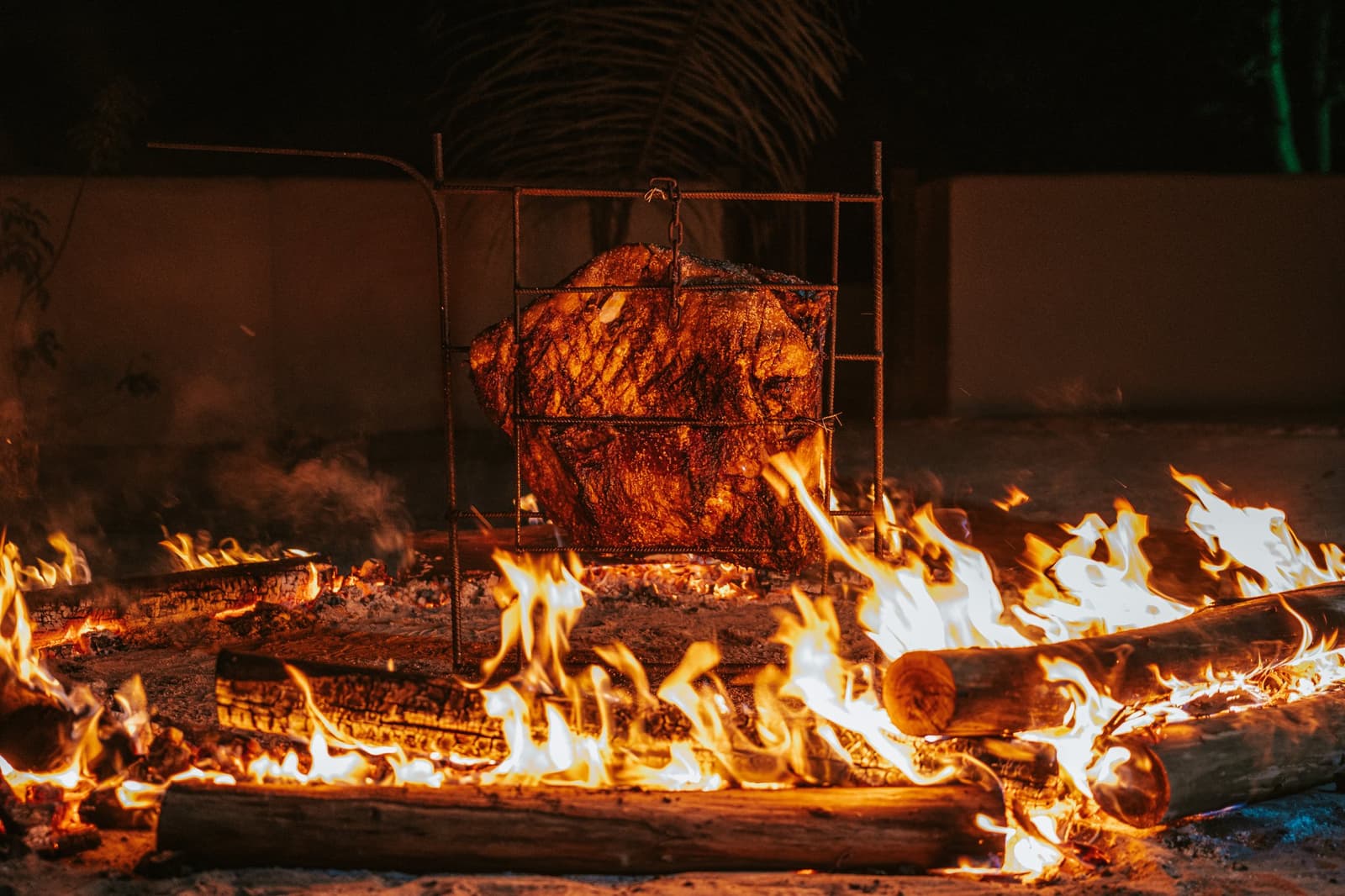 Slow-roasted over real wood, the way patience pays off at the table. A large cut of meat slow-roasting in a rustic iron cage grill over open wood flames at night at Pousada Trijunção, deep in the Cerrado