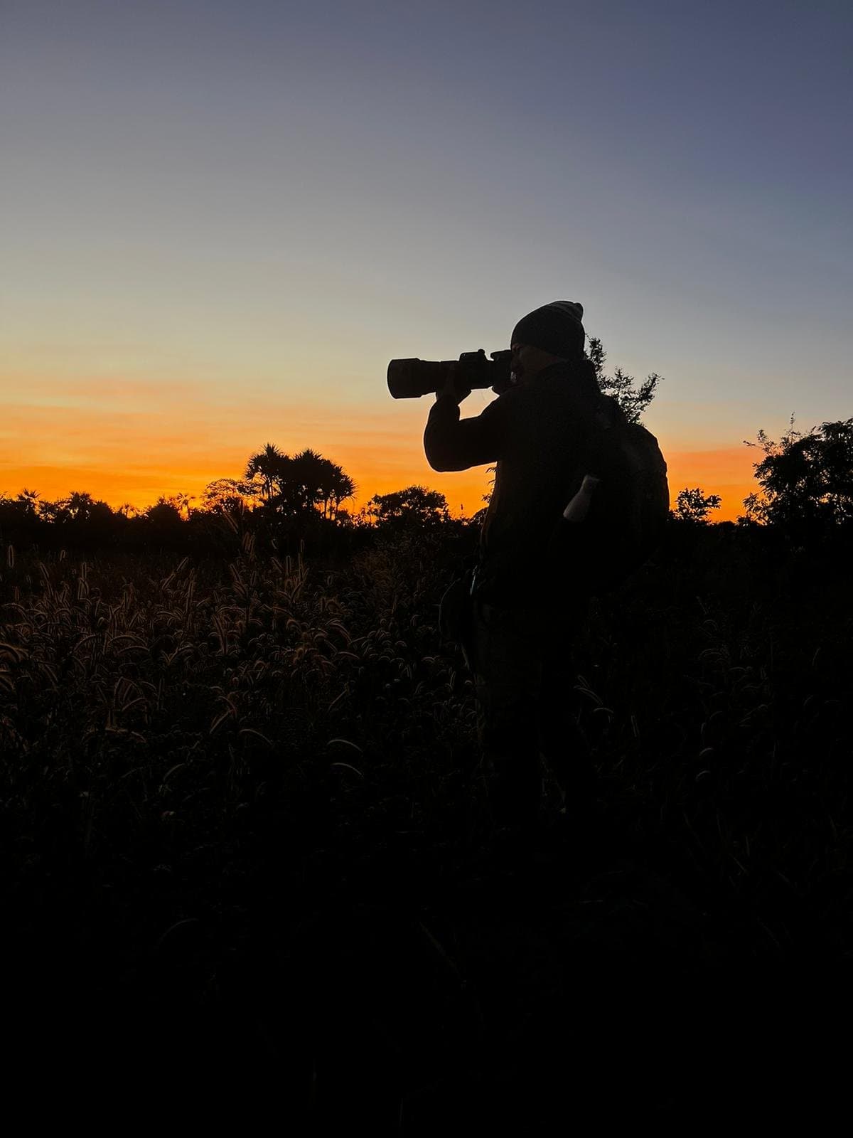 The light only lasts a few minutes. Good thing someone's ready. Silhouette of a wildlife photographer with a telephoto lens raised to their eye, standing among tall Cerrado grasses against a vivid orange and blue sunrise sky at Pousada Trijunção