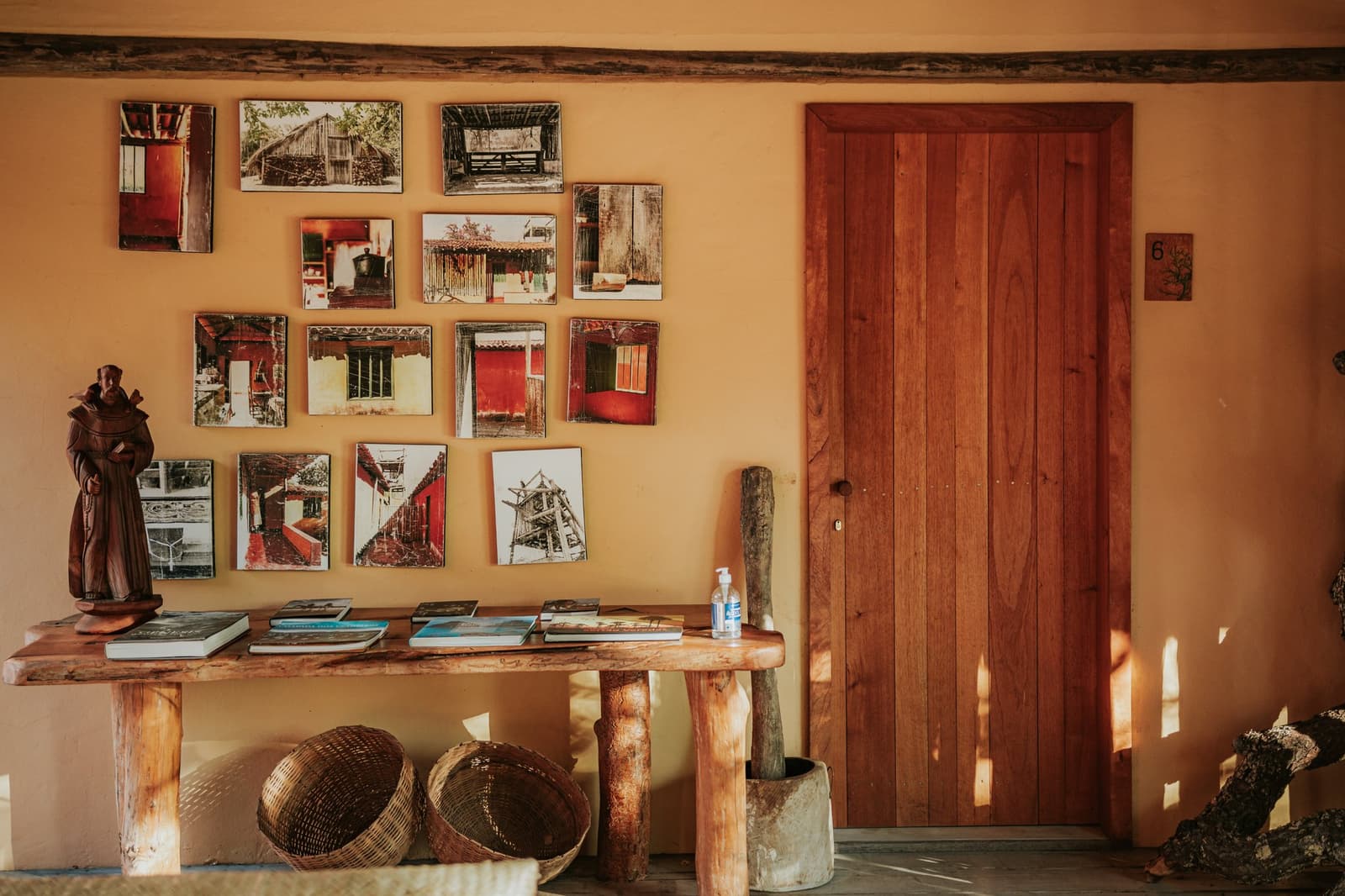 Every photo on that wall has a story the staff will happily tell you. Warm ochre wall at Pousada Trijunção decorated with a grid of photographic prints depicting traditional Cerrado architecture, beside a handcrafted wooden console table holding books, a carved monk figurine and woven baskets