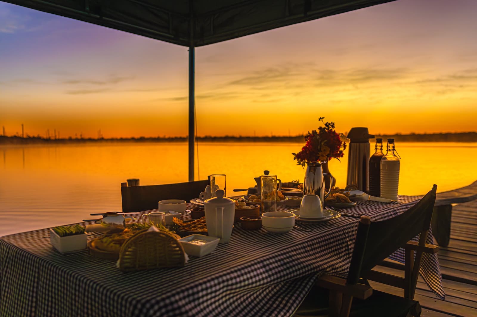 The kind of morning that makes you forget what time zone you're in. A fully set breakfast table on a wooden pier at Pousada Trijunção, with a gingham tablecloth, fresh flowers, coffee pots and fruit, silhouetted against a vivid golden sunrise over a glassy lake