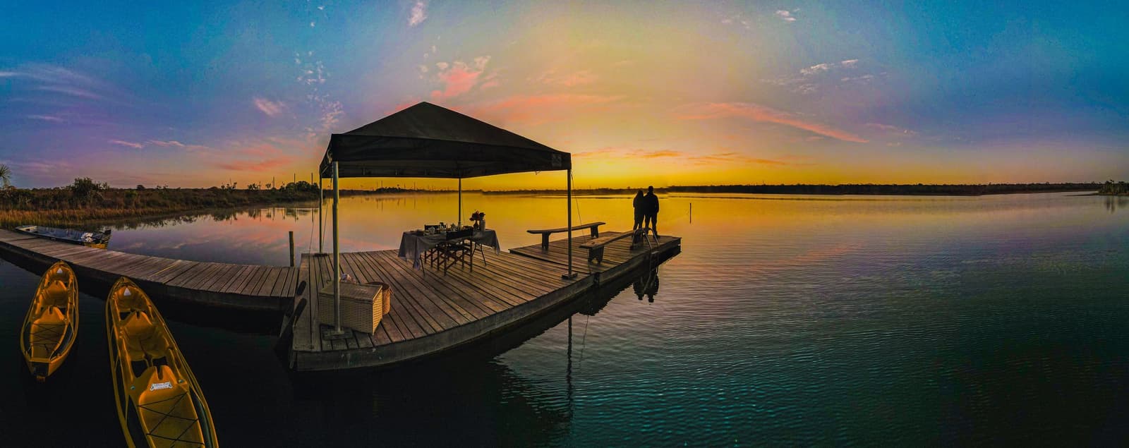 Dinner on the dock, the whole wetland sky to yourselves. A couple silhouetted at the end of a wooden pier at Pousada Trijunção, a canopied breakfast table behind them and kayaks moored to the left, as a golden sunrise blazes across the glassy lake