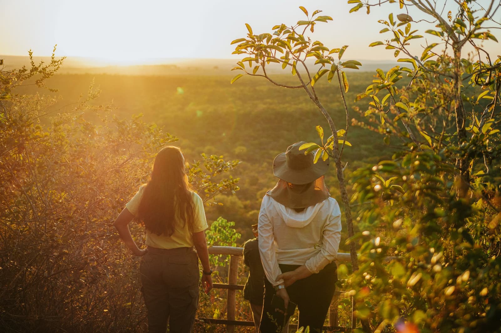Some sunsets you just have to stop talking and watch. Two women standing at a wooden railing viewpoint at Pousada Trijunção, gazing over a vast golden-lit Cerrado landscape at sunset