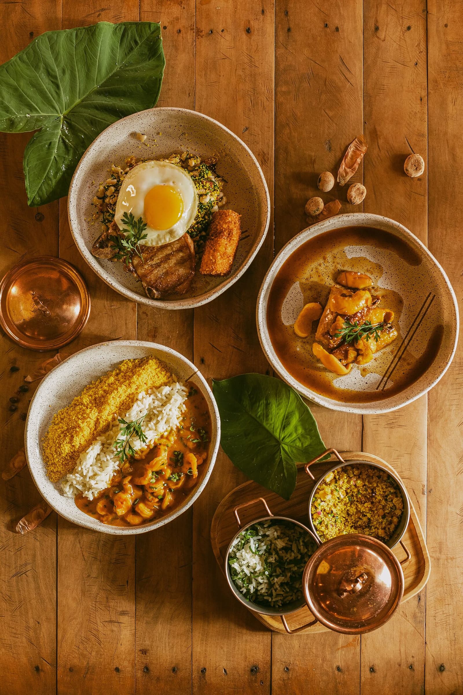 The kind of lunch that makes you forget you had a plan for the afternoon. Four ceramic bowls of traditional Brazilian dishes served at Pousada Trijunção, including shrimp moqueca with rice and farofa, a fried-egg rice bowl with crispy pork belly, a rich meat stew, and copper pots of sides on a warm wooden table