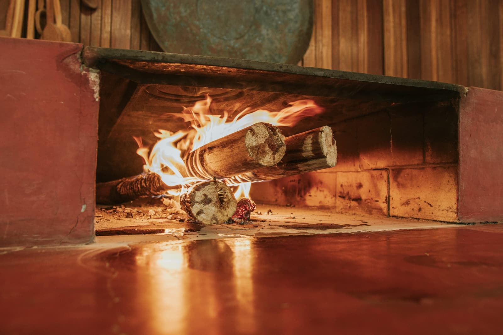 Good food starts here, long before anything reaches the table. Thick wooden logs burning with open flames inside a traditional wood-fired stone hearth at Pousada Trijunção, with warm firelight reflecting off the polished red floor