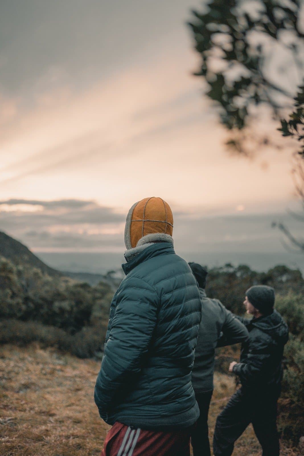 Pack an extra layer, mornings up here have opinions. Two hikers in heavy winter layers and a sheepskin hat stand on an exposed ridge during Serra Fina Trekking, gazing over misty mountain valleys beneath a soft pastel sunrise sky in the Serra da Mantiqueira