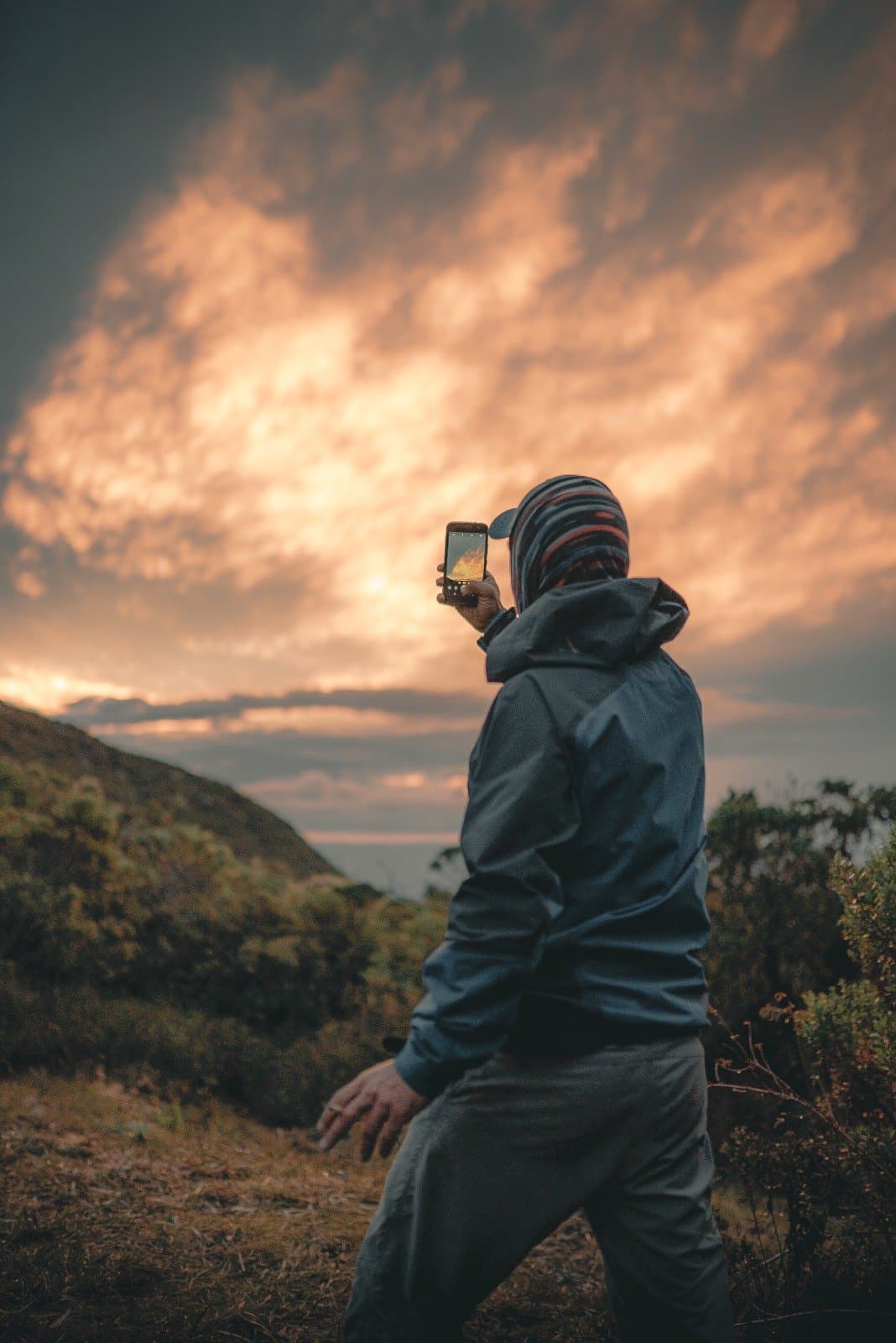 Some skies demand you stop walking and just point your phone. A hiker in a jacket and striped beanie photographs a dramatic orange and gold sunset sky above the Serra da Mantiqueira ridgeline during a Serra Fina Trekking expedition
