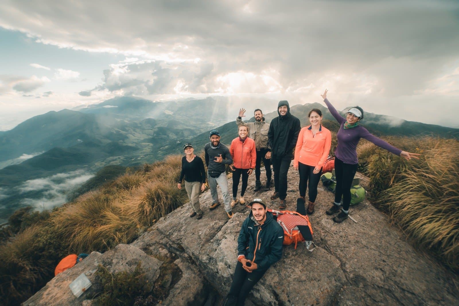 That collective exhale at the top, worth every steep step to get there. Eight hikers celebrate on a rocky summit during the Serra Fina Trekking, with dramatic cloud-filled skies and the rolling ridges of the Serra da Mantiqueira stretching to the horizon