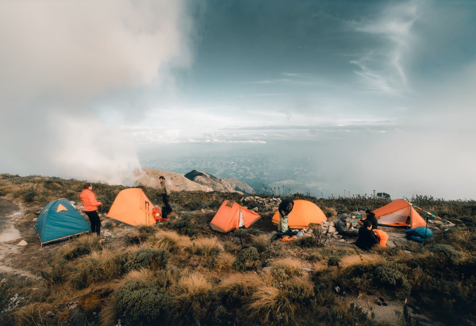 Camp is set, clouds are moving in, and nobody's complaining. A group of trekkers setting up orange and teal tents at a Serra Fina Trekking high-altitude wild camp on an exposed ridge in the Serra da Mantiqueira, with sweeping views of valleys and dramatic storm clouds above