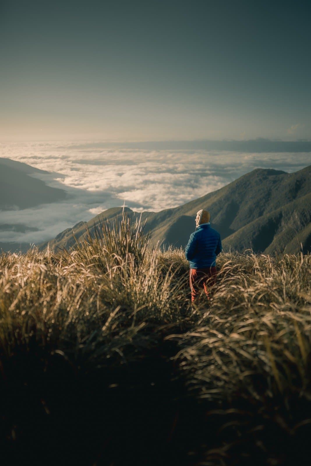 Up here, the clouds are the floor and the sky is all yours. A hiker in a blue down jacket stands among golden highland grasses on the Serra Fina Trekking route, gazing over a vast sea of clouds filling the valleys of the Serra da Mantiqueira