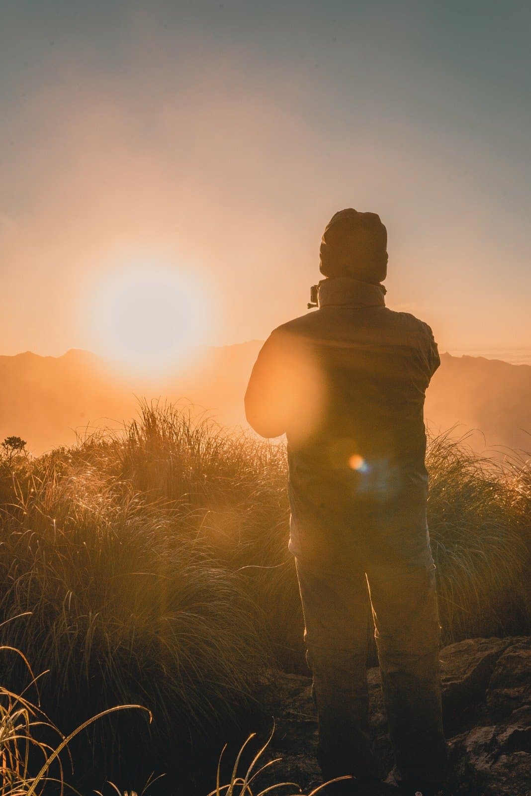 First light on the ridge, and suddenly the early wake-up makes perfect sense. A hiker's silhouette stands among golden mountain grasses at sunrise during a Serra Fina Trekking expedition, with misty ridges of the Serra da Mantiqueira glowing orange in the background