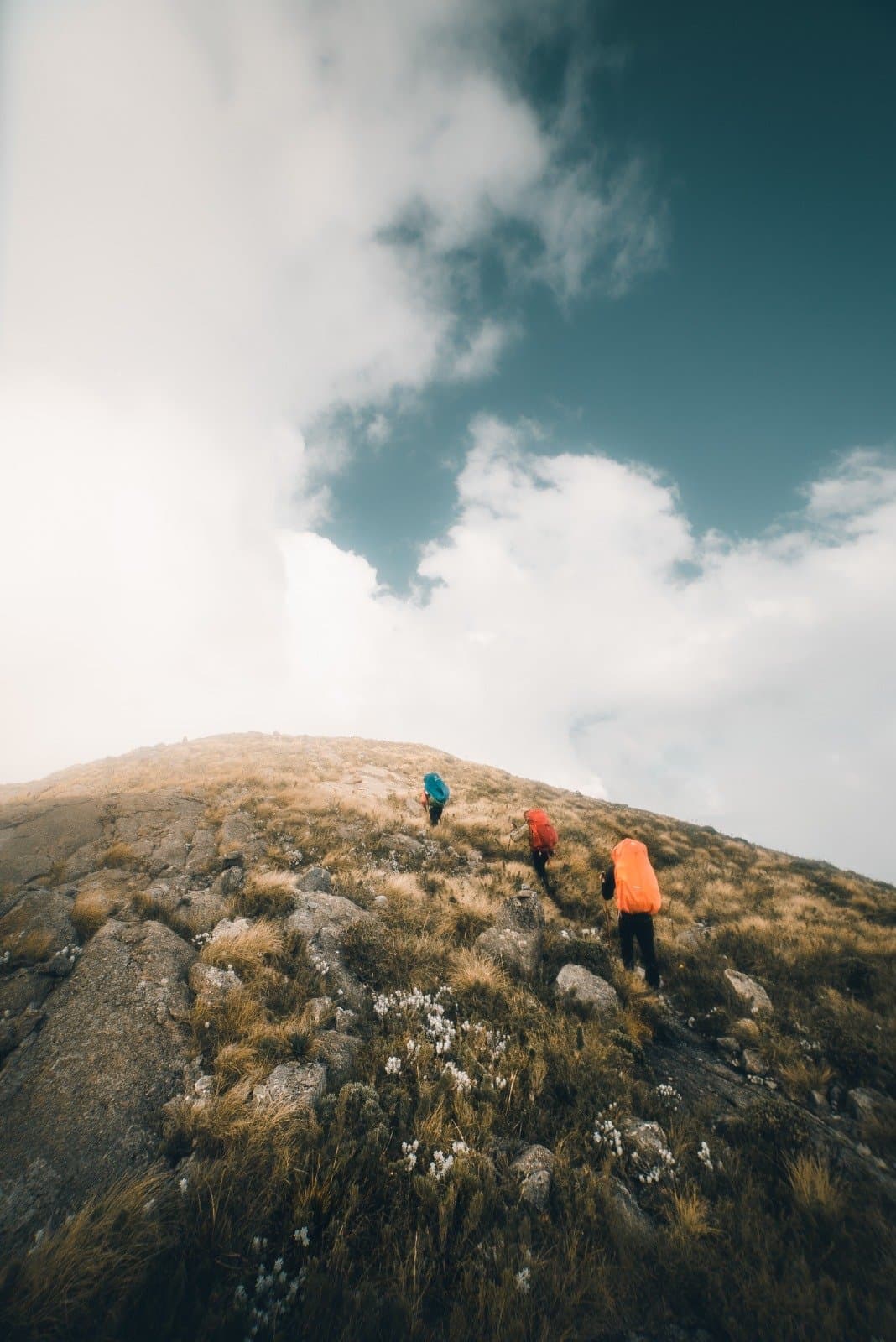 The summit won't come to you, so you go to it. Three hikers with colourful backpacks ascending a steep rocky ridge on the Serra Fina Trekking route in the Serra da Mantiqueira, under a dramatic sky