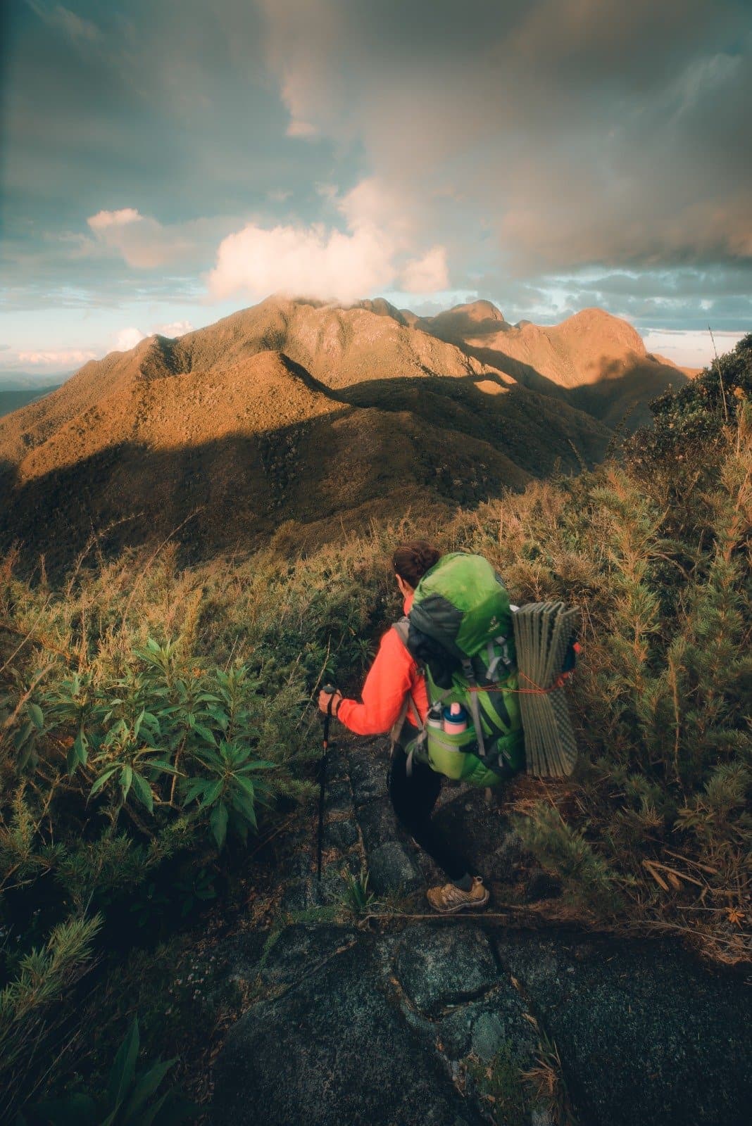 One foot in front of the other, the Serra Fina does the rest. A backpacker in an orange jacket carrying a full pack pauses on a rocky ridge during Serra Fina Trekking, with golden-lit mountain summits of the Serra da Mantiqueira stretching under a dramatic cloudy sky