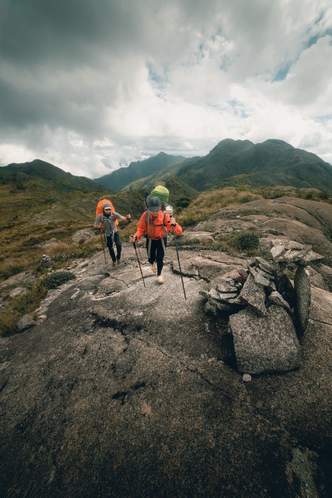 Granite underfoot, clouds overhead, and a long way still to go. Two backpackers in orange jackets using trekking poles to navigate a granite rock slab on the Serra Fina Trekking route, with green mountain ridges and dramatic clouds filling the Serra da Mantiqueira skyline behind them