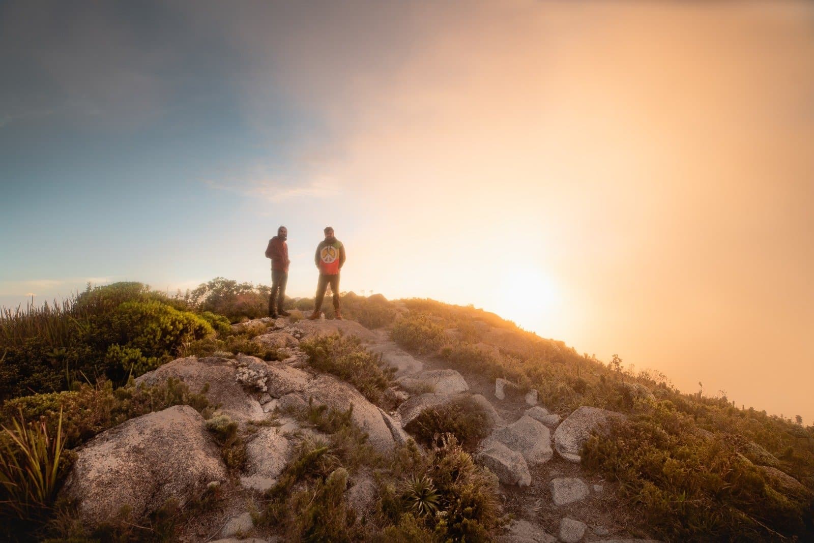 The summit doesn't care what time you set your alarm, it rewards every one. Two hikers silhouetted against a glowing golden sunrise on a rocky Serra Fina Trekking summit, surrounded by high-altitude Atlantic forest vegetation in the Serra da Mantiqueira.