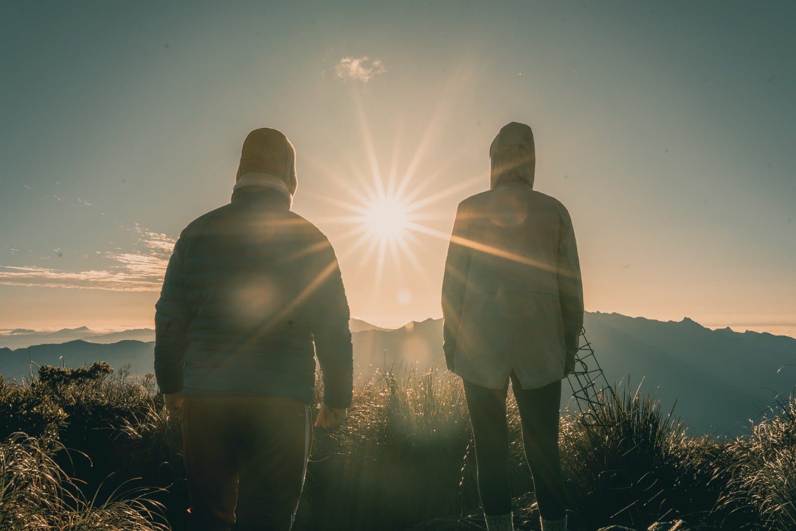 Some moments you just stand still and let the mountain do the talking. Two hooded hikers silhouetted against a brilliant starburst sunrise on a Serra Fina Trekking summit, with layers of Mantiqueira mountain ridges stretching to the horizon