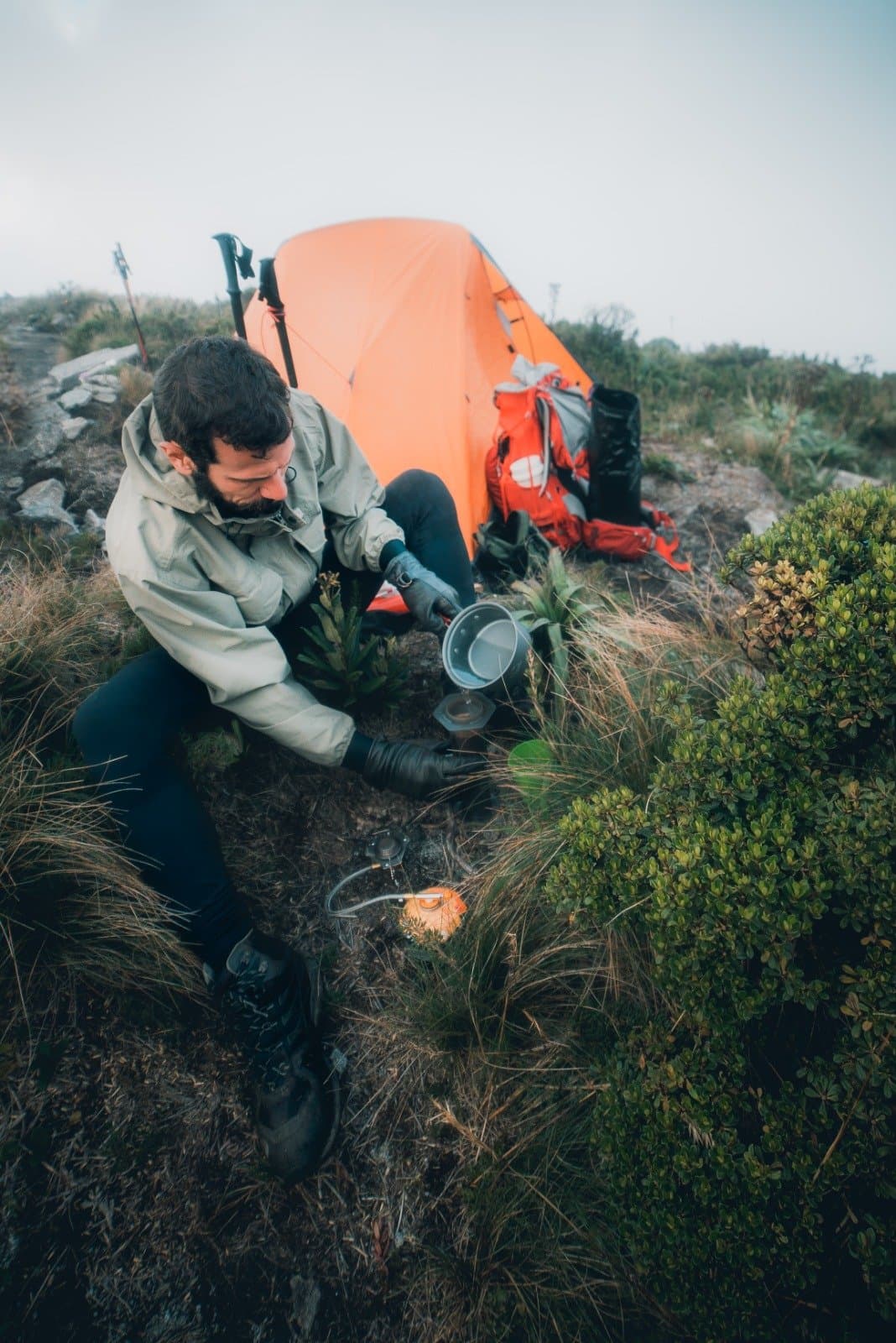 Hot food hits different at altitude, in the mist, with cold hands. A bearded hiker in a rain jacket pours water into a pot on a compact camp stove beside an orange tent and red backpack during a Serra Fina Trekking expedition in the Serra da Mantiqueira highlands