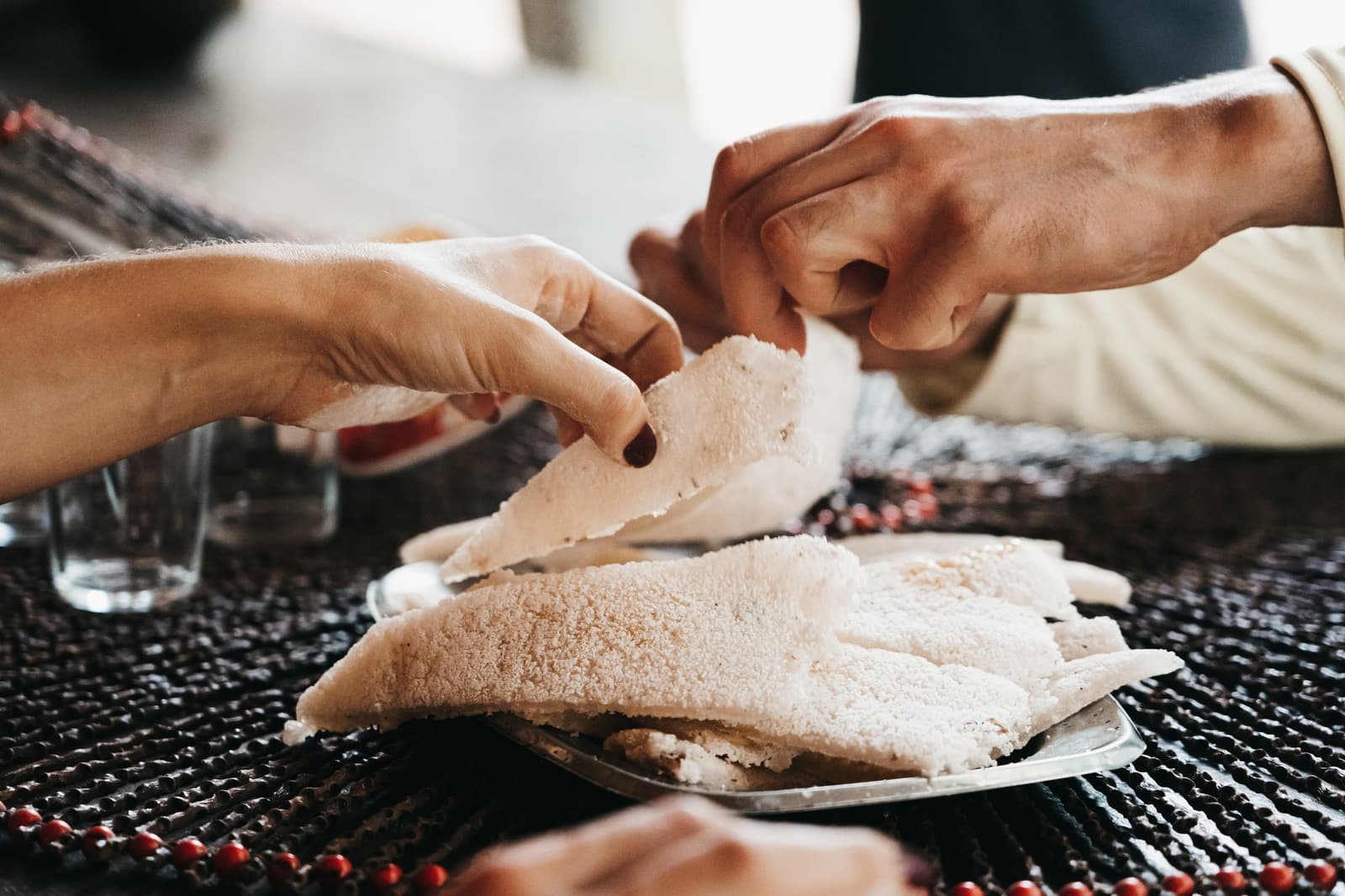 Tapioca tastes better torn, not cut. Ask anyone along the river. Two people breaking apart a piece of soft white cassava flatbread over a shared plate on an untamed amazon cruise meal table