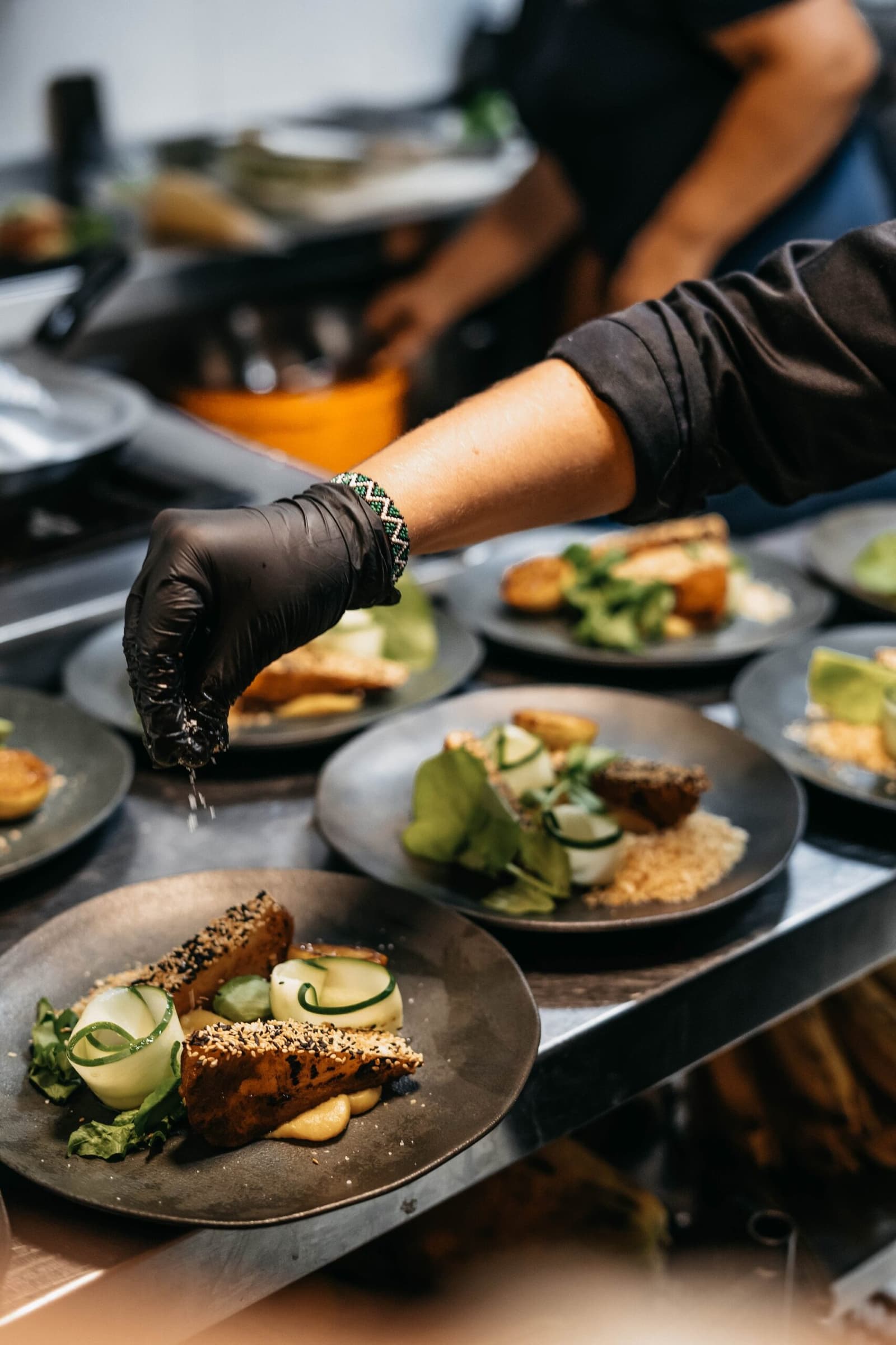 Good food doesn't take a day off, not even on the river. A chef wearing black gloves sprinkles seasoning over multiple elegantly plated dishes featuring sesame-crusted portions, spiralled cucumber and fresh greens, prepared aboard the untamed amazon cruise