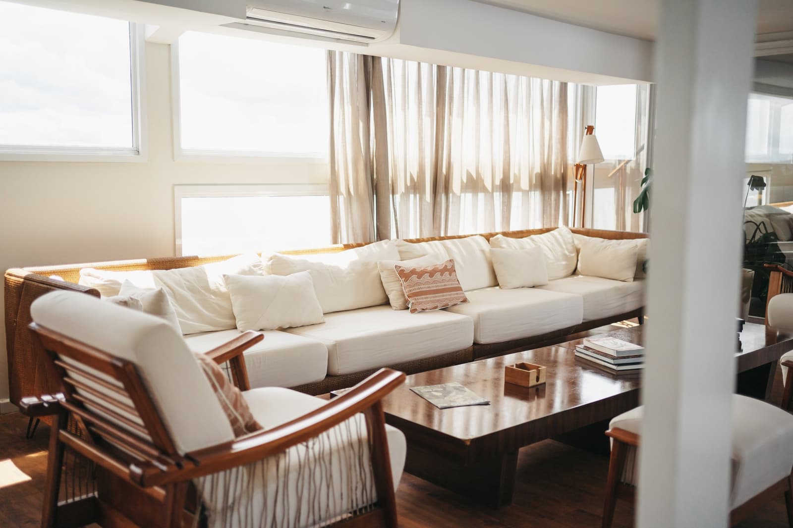 A room that earns its keep between river landings. Sunlit social lounge on the untamed amazon cruise featuring a wide cream sofa with rattan frame, wooden armchairs, and a polished coffee table beneath sheer linen curtains