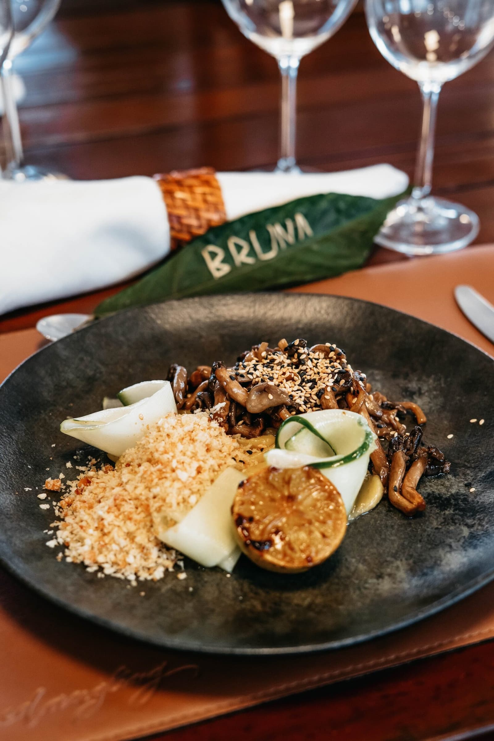 Dinner on the river, where the chef clearly has something to prove. A sautéed mixed mushroom dish with sesame seeds, pickled vegetables, toasted breadcrumbs and a caramelised round vegetable slice, plated on a dark stone dish aboard the untamed amazon cruise, with a woven-ring napkin and a leaf place card reading 'Bruna' in the background
