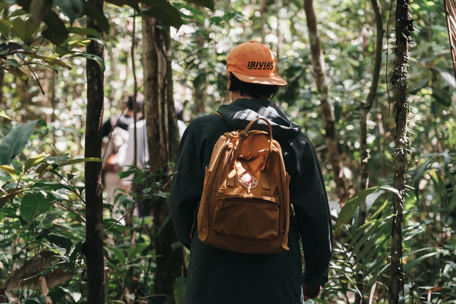 The forest does the leading, you just follow. A hiker with an orange cap and tan backpack walks through dense rainforest undergrowth on an untamed amazon cruise excursion, with fellow travellers visible ahead on the trail