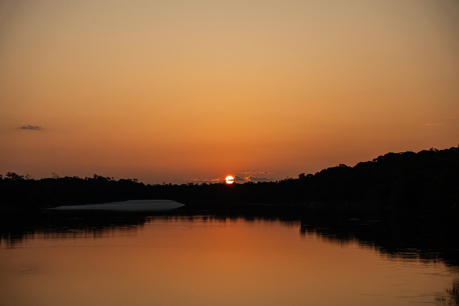 The river holds the whole sky at the end of the day. A glowing orange sun sinks behind a dark treeline silhouette on an untamed Amazon cruise, its reflection shimmering across a calm river with a white sandbar visible on the left bank