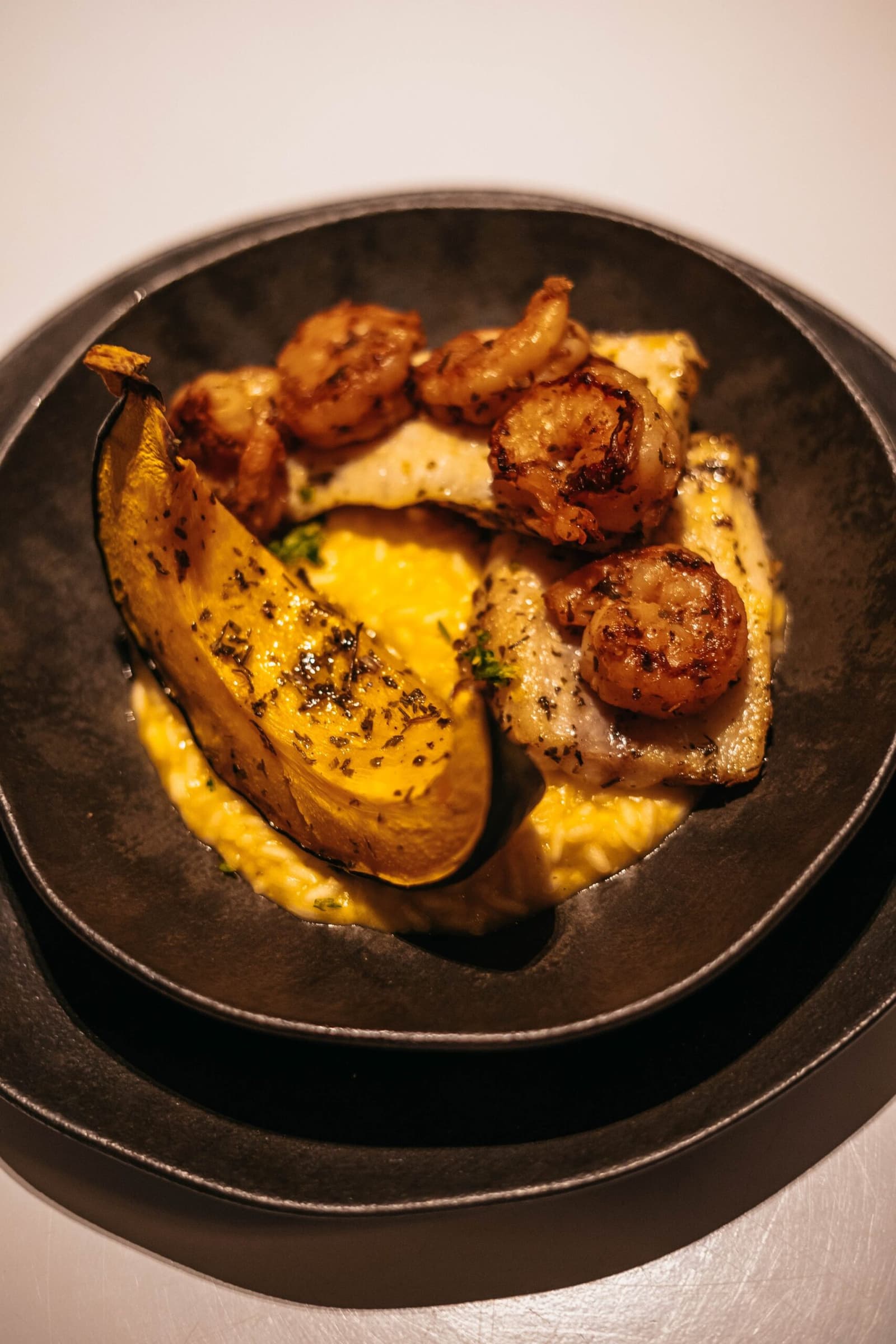River fish, shrimp, and roasted plantain, the Amazon on a plate. Seared shrimp and white fish fillet plated on a golden cream sauce with a charred plantain half, served in a matte black bowl on the untamed amazon cruise