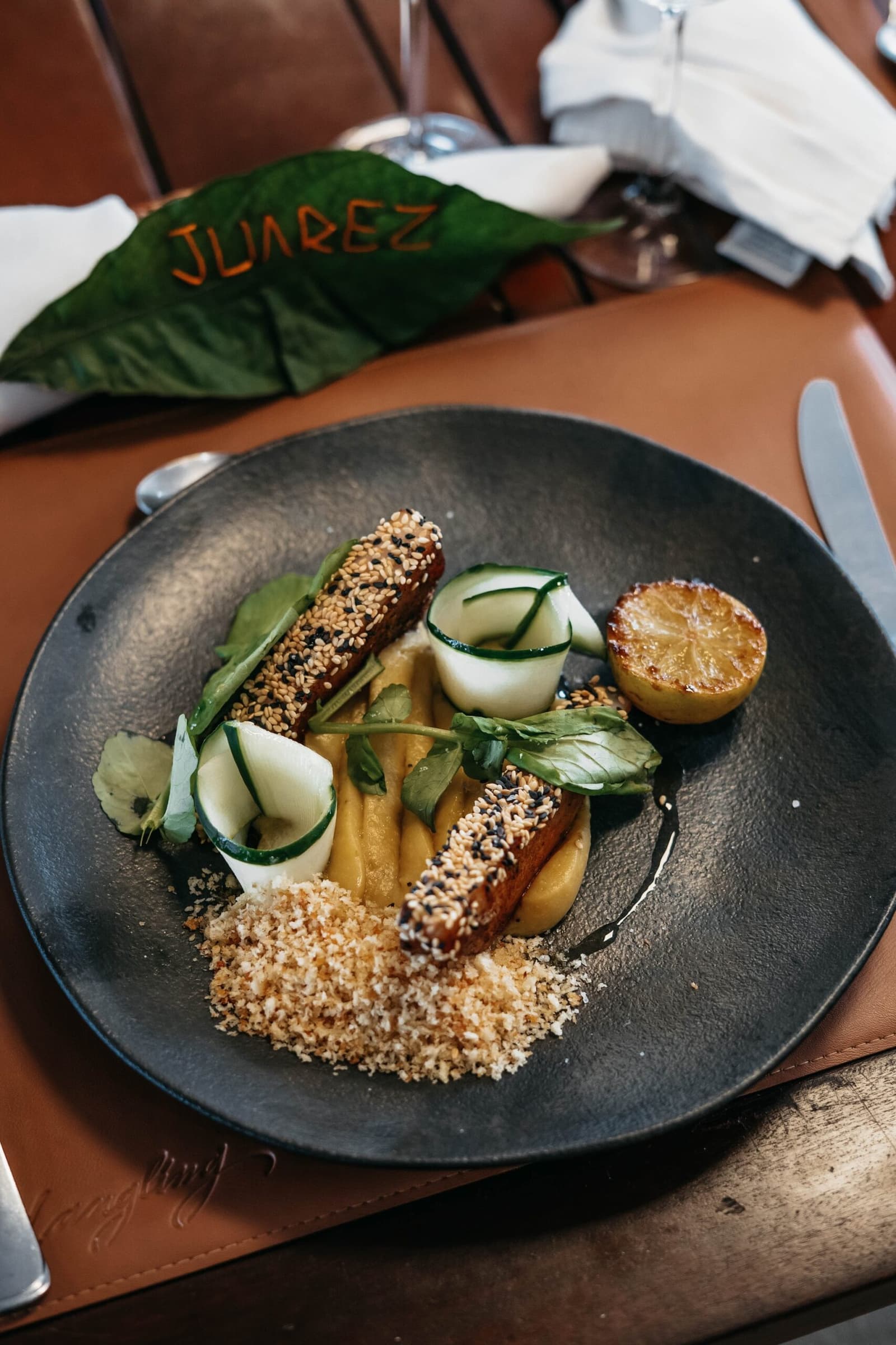 Juarez knows a leaf makes the best business card. Sesame-crusted protein strips with cucumber rolls, golden purée, breadcrumbs and a charred citrus half plated on a dark slate dish aboard the untamed amazon cruise, with a leaf branded 'Juarez' in the background