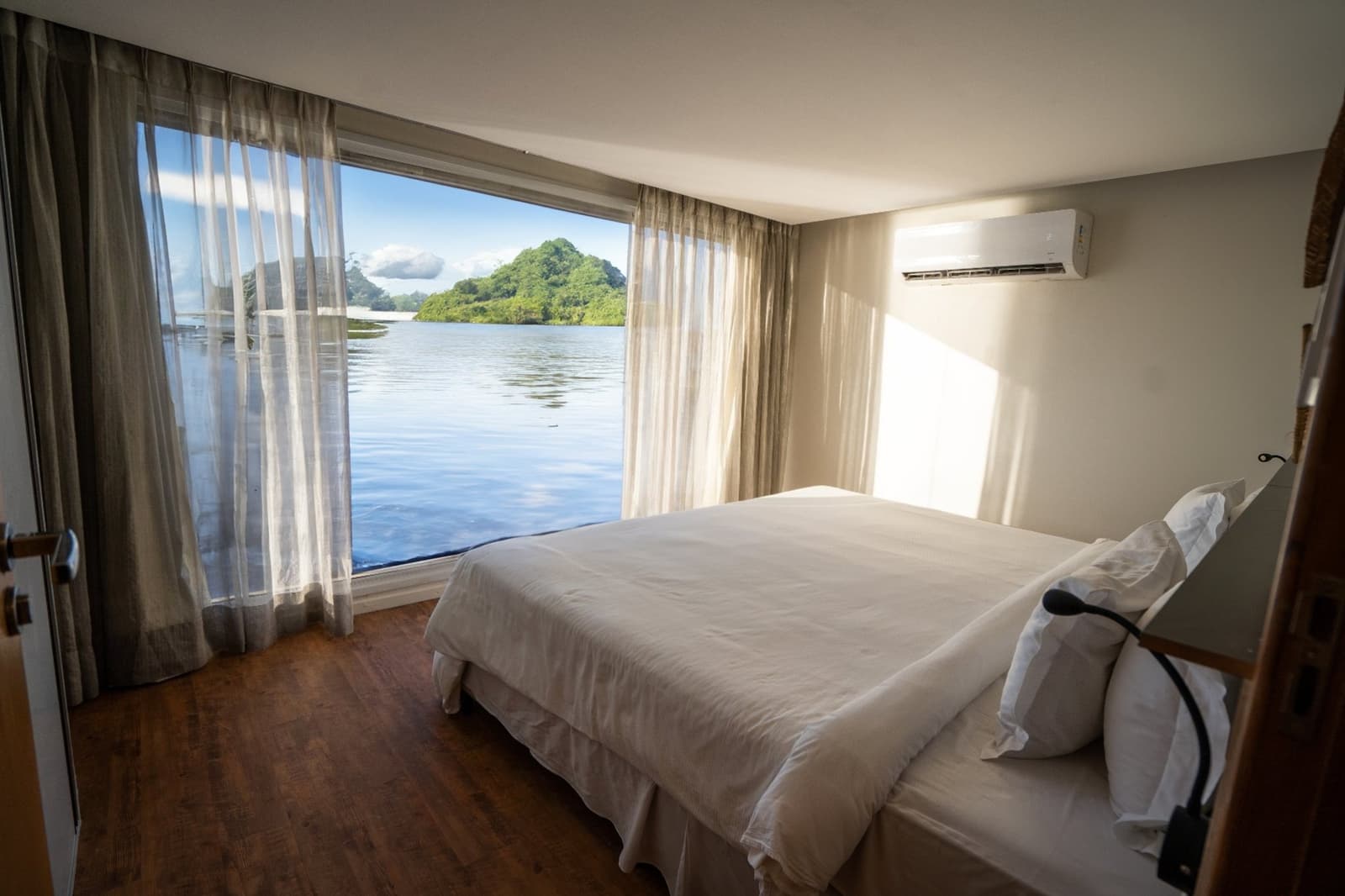 Your morning view, sorted. The river does the rest. A bright, air-conditioned cabin on the untamed amazon cruise featuring a double bed with white linen, hardwood floors, and a floor-to-ceiling window overlooking a calm river and tree-covered island