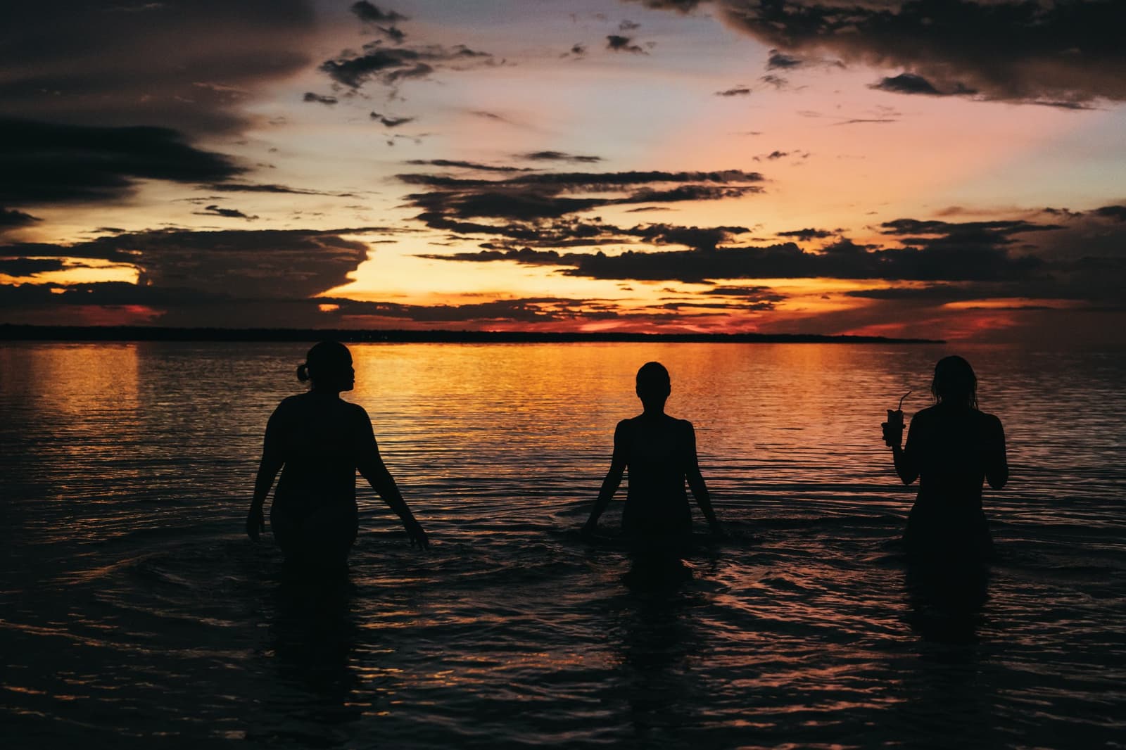 The river keeps the day going a little longer than the sky does. Three guests silhouetted waist-deep in calm river water during a vivid orange and red sunset on an untamed amazon cruise