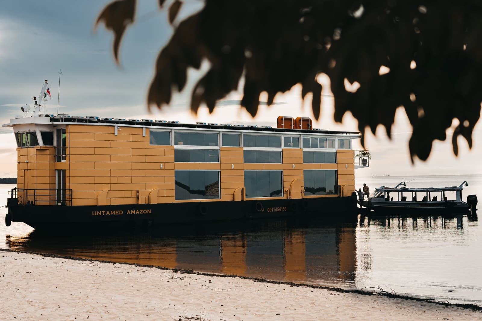 Your floating base camp, ready and waiting on the river. The Untamed Amazon cruise vessel moored at a sandy riverbank at sunset, its yellow hull and panoramic cabin windows reflected in the calm water, with a smaller excursion boat alongside