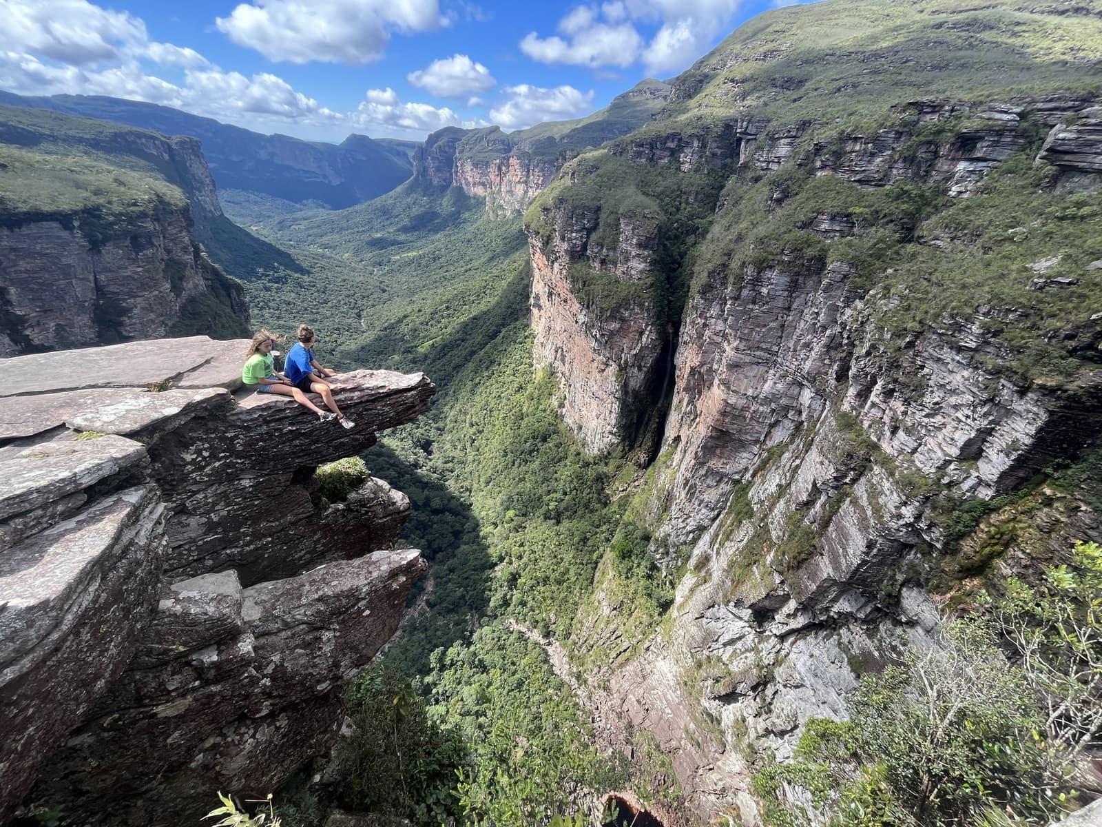 The kind of lunch spot that makes you forget you have legs. Two hikers sitting on a rock ledge overlooking the deep canyon and lush green valley of the Vale do Pati Trek in Chapada Diamantina