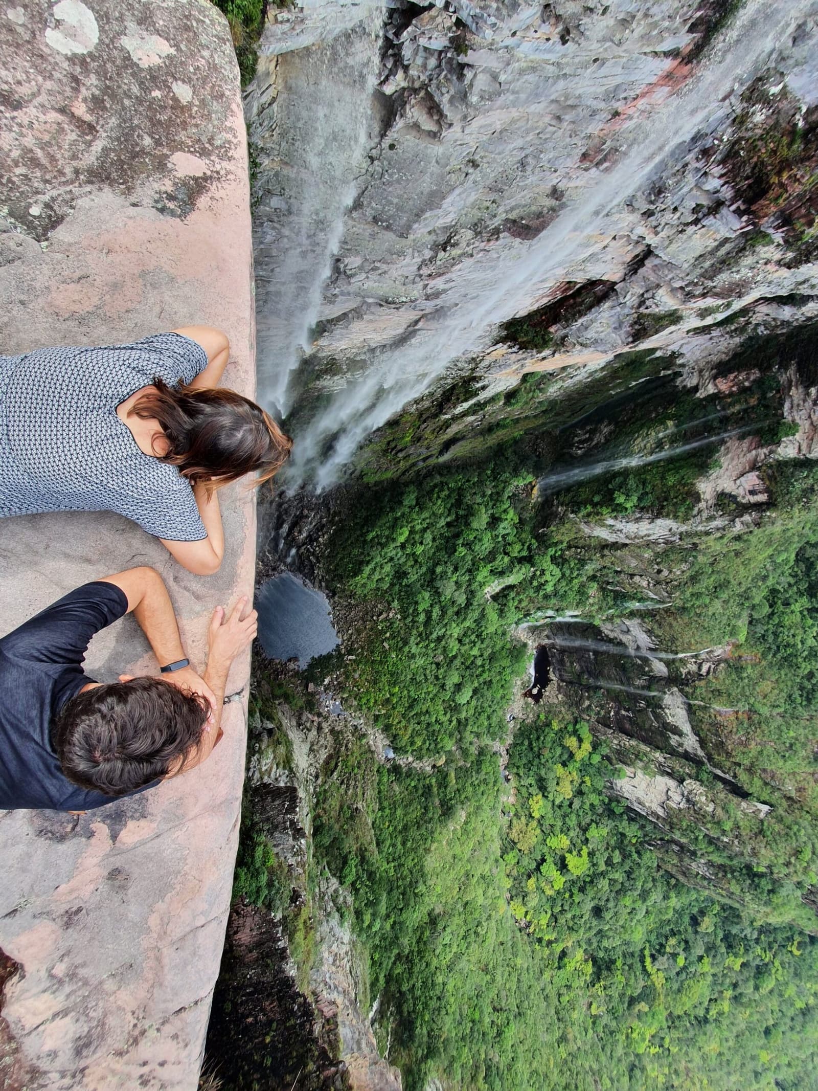 The drop is very real, and so is the grin right after. Two hikers lie flat on a smooth sandstone ledge peering straight down a towering cliff face on the Vale do Pati Trek, with a thin waterfall and dense green canyon far below