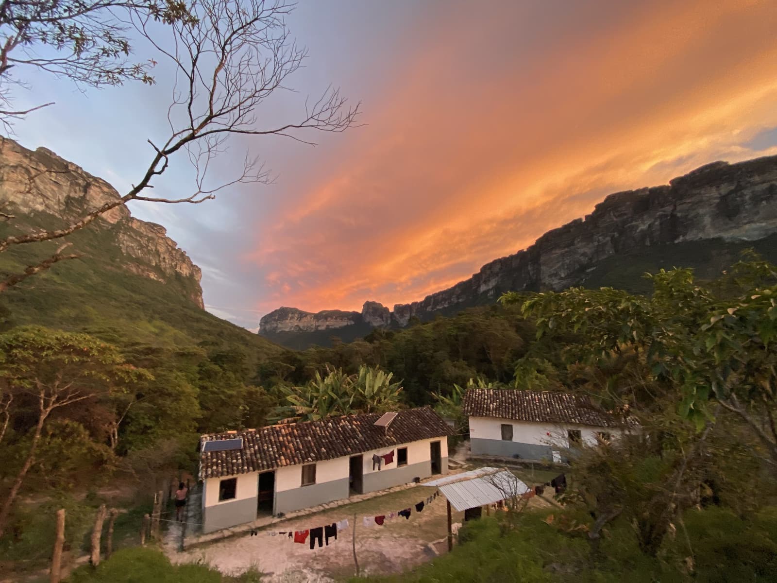 End of a long day on the trail, and the valley puts on a show. A rustic white farmhouse with terracotta roof tiles and laundry drying on a line, nestled in a valley between dramatic sandstone cliffs under a blazing orange and pink sunset sky on the Vale do Pati Trek