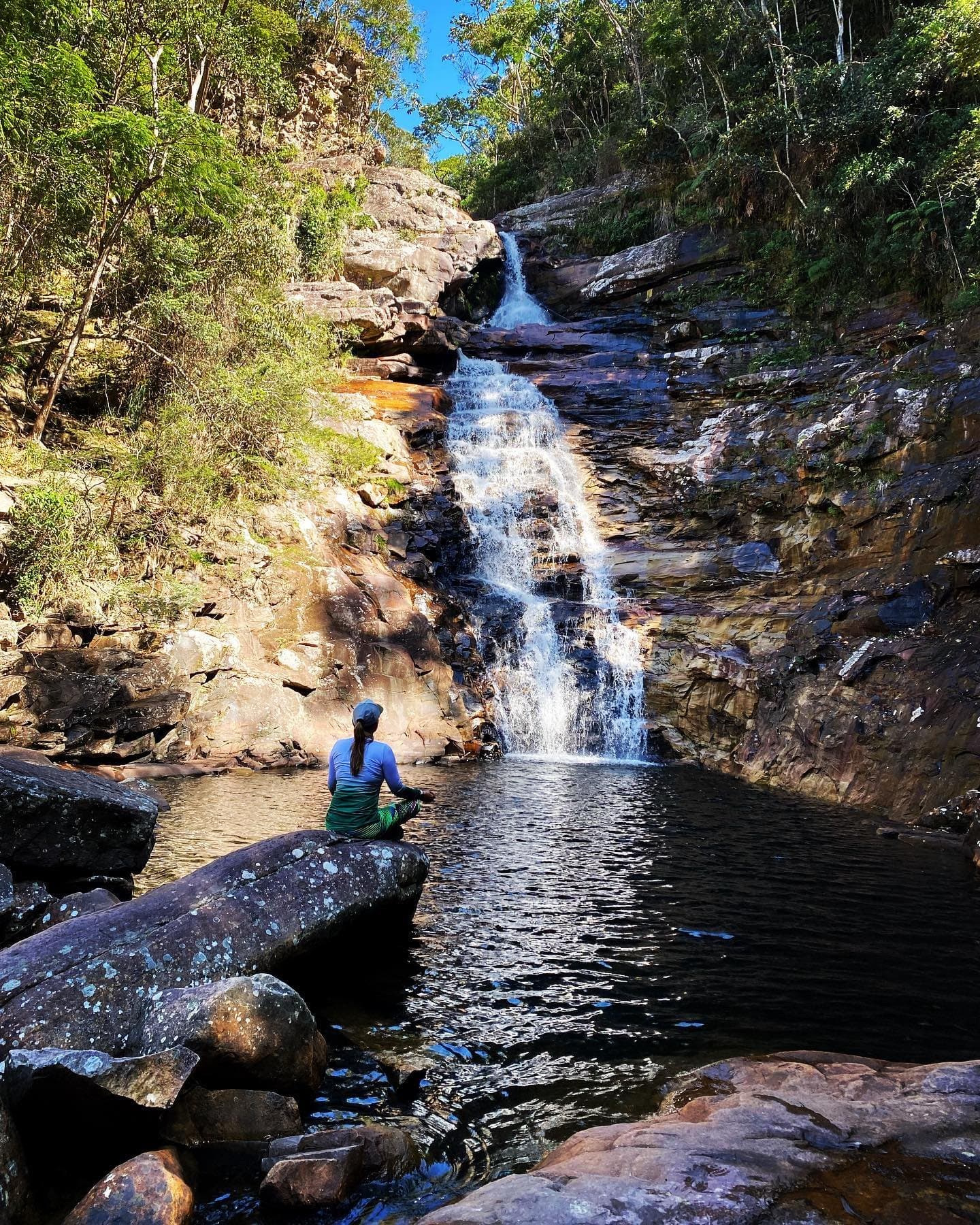 Find a rock, sit down, let the waterfall do the talking. A hiker sits on a rock beside the natural pool of a tiered waterfall cascading down iron-streaked sandstone cliffs on the Vale do Pati Trek, surrounded by green Atlantic forest under a blue sky
