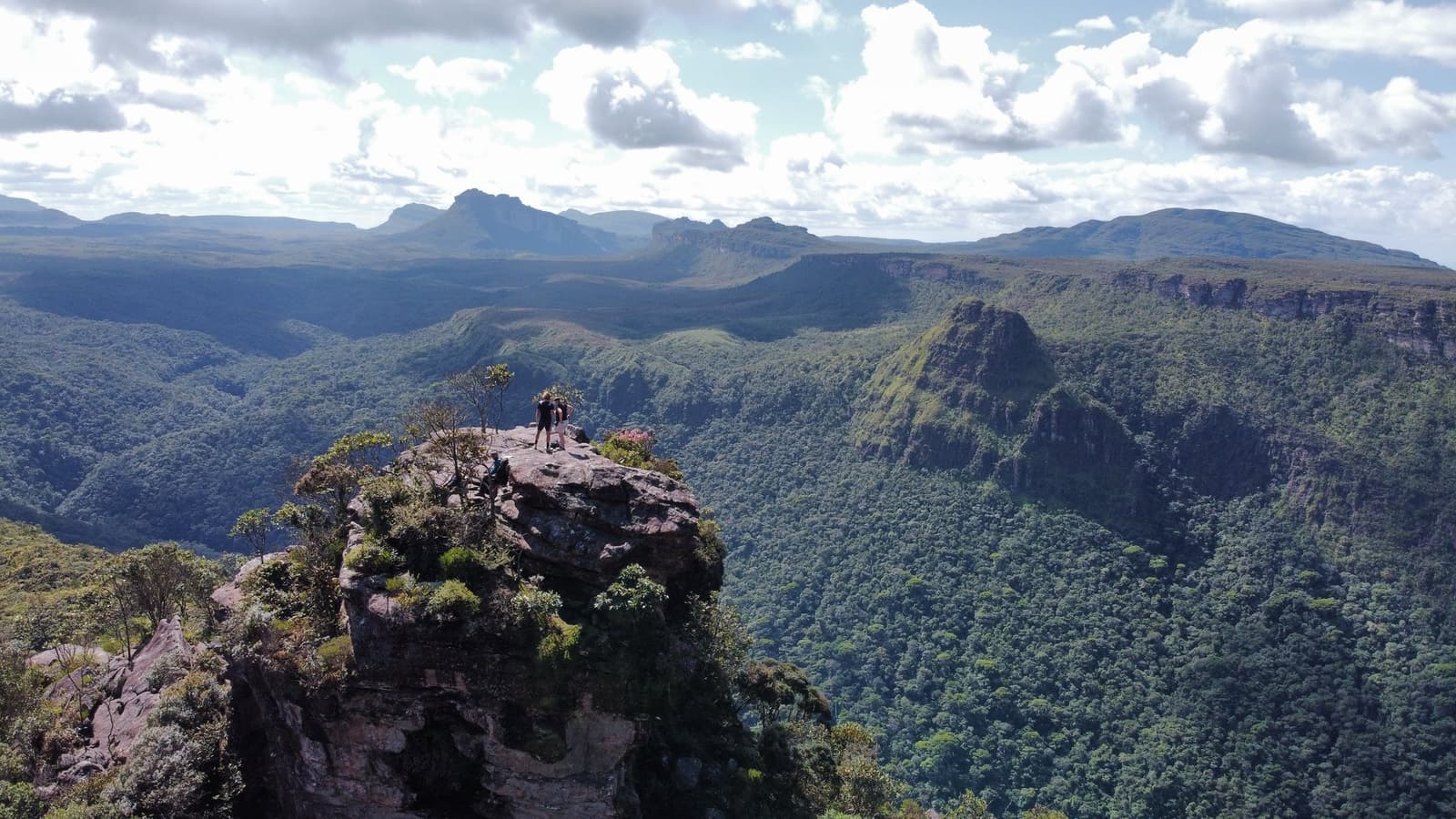 That ledge earns every step of the Vale do Pati trail. Two hikers stand on a rocky outcrop high above a forested canyon on the Vale do Pati Trek, with layered sandstone plateaus and green valleys stretching to the horizon under a partly cloudy sky