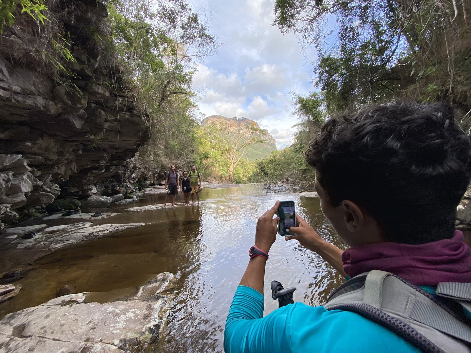 Someone's got to document the moment, might as well be you. A hiker photographs two smiling trekkers standing in a shallow rocky river gorge on the Vale do Pati Trek, with sandstone cliffs and a mesa visible in the background