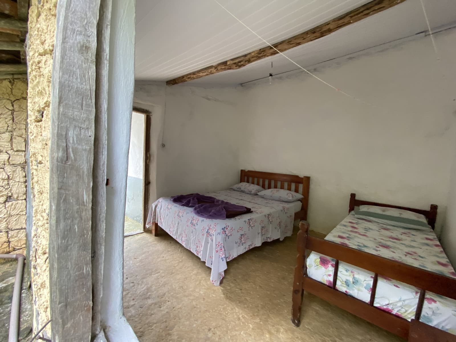 No luxury here, just honest rest after a long day on the trail. Simple shared bedroom in a Vale do Pati Trek family homestay, with two wooden-framed beds covered in floral bedspreads, exposed ceiling beam, and stone-and-plaster walls