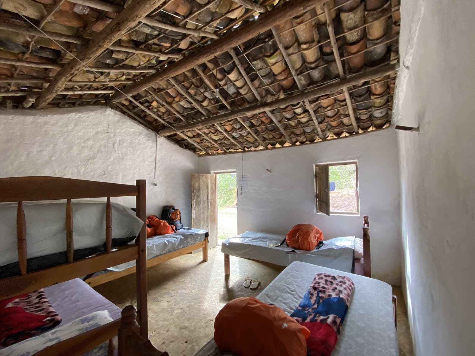 No frills, but the clay-tile ceiling has more character than most hotel rooms. Simple shared bedroom in a Vale do Pati Trek family homestay, with wooden bunk beds and single beds topped with orange dry bags, exposed clay-tile roof on timber beams, and whitewashed walls