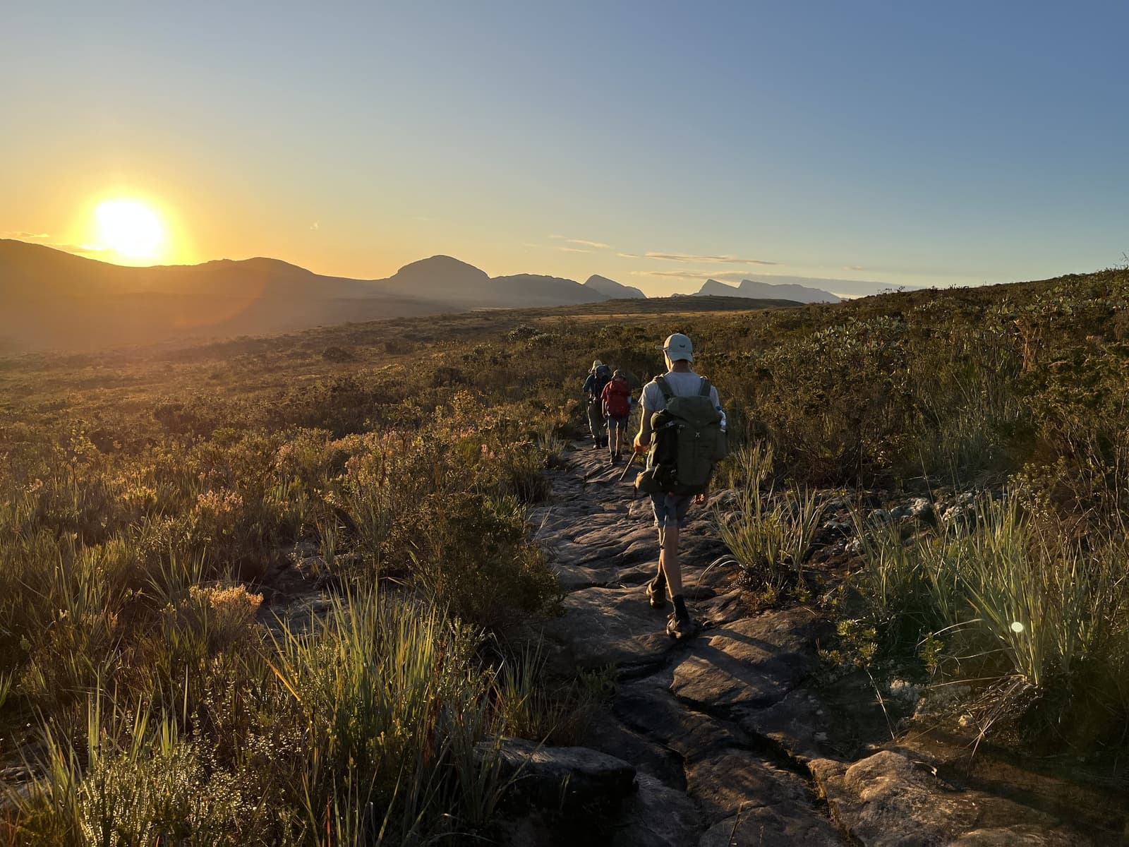 First light on the trail, and the day already feels earned. Three hikers walking a rocky trail through campo rupestre scrubland on the Vale do Pati Trek as a large golden sun sets over a distant mountain ridge