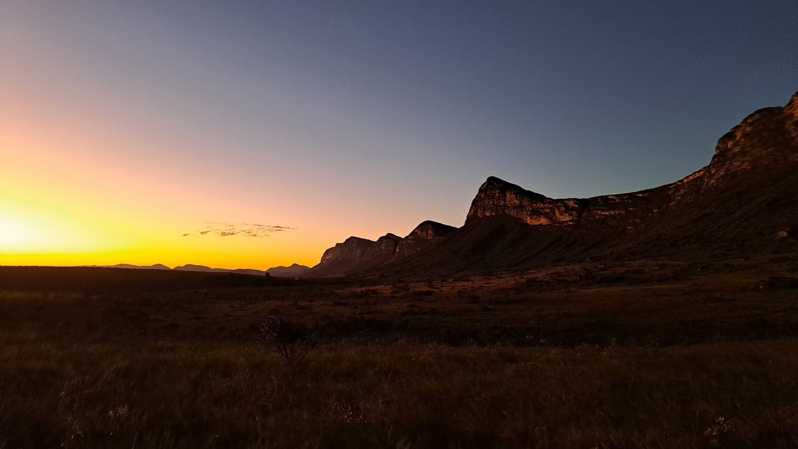 The light clocks out early here, and the rocks make it count. Fiery orange and gold sunset over a vast open savanna with a rugged rocky escarpment silhouetted against a deep blue sky on the Vale do Pati Trek
