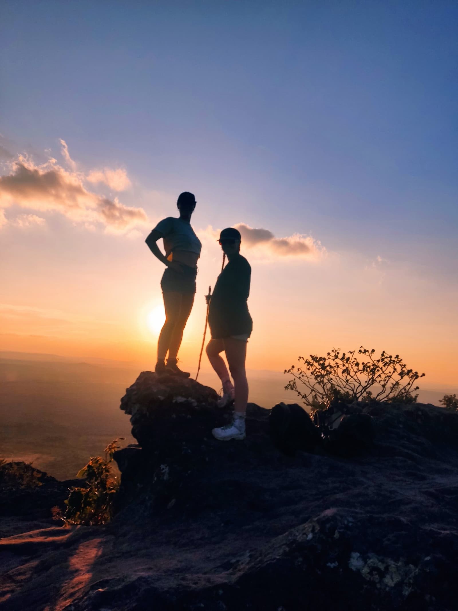 The summit earns its keep right about now. Two hikers silhouetted on a rocky summit at sunset during the Vale do Pati Trek, with a vast lowland panorama glowing orange beneath a wide sky