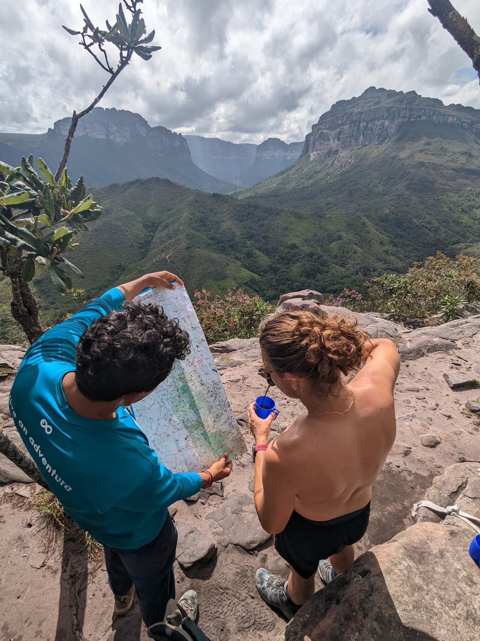 The map knows the way. Your guide makes sure you do too. A guide in a teal shirt and a hiker study a trail map together on a rocky outcrop during the Vale do Pati Trek, with dramatic sandstone tepuis and a lush green valley stretching behind them under a cloudy sky