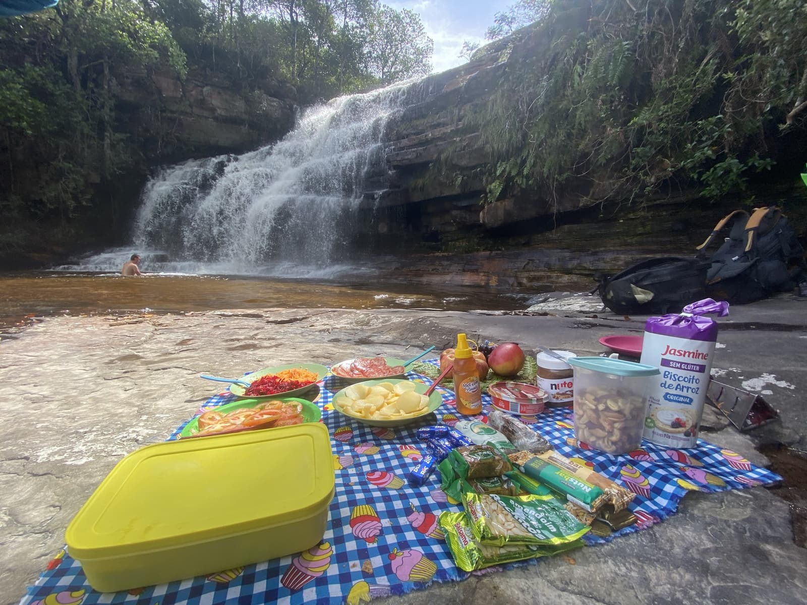 Lunch with a better view than most restaurants will ever manage. A colorful trail picnic laid out on a checkered blanket over flat rocks beside a cascading waterfall on the Vale do Pati Trek, with salads, fruit, nuts, honey, and snacks, while a swimmer enjoys the natural pool in the background
