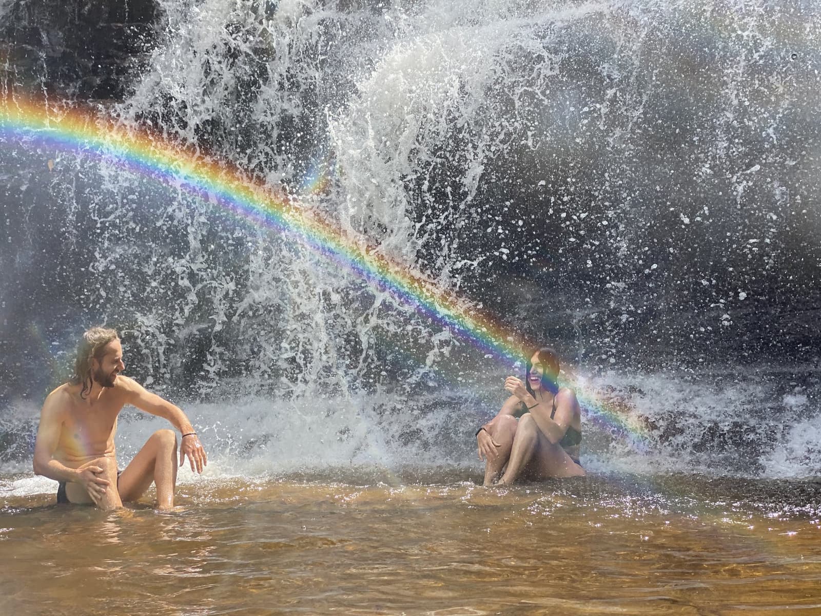 A rainbow appeared right on cue. No filter needed. Two smiling travelers sitting in a shallow natural pool at the base of a cascading waterfall on the Vale do Pati Trek, a vivid rainbow arching through the mist between them