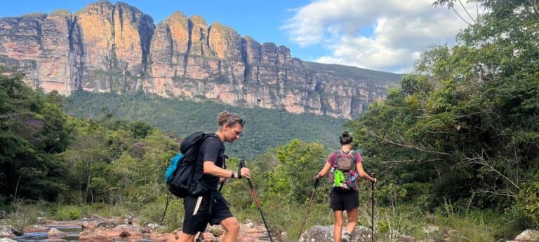 Two trekkers with backpacks and trekking poles crossing a rocky riverbed below the cliff walls of Chapada Diamantina