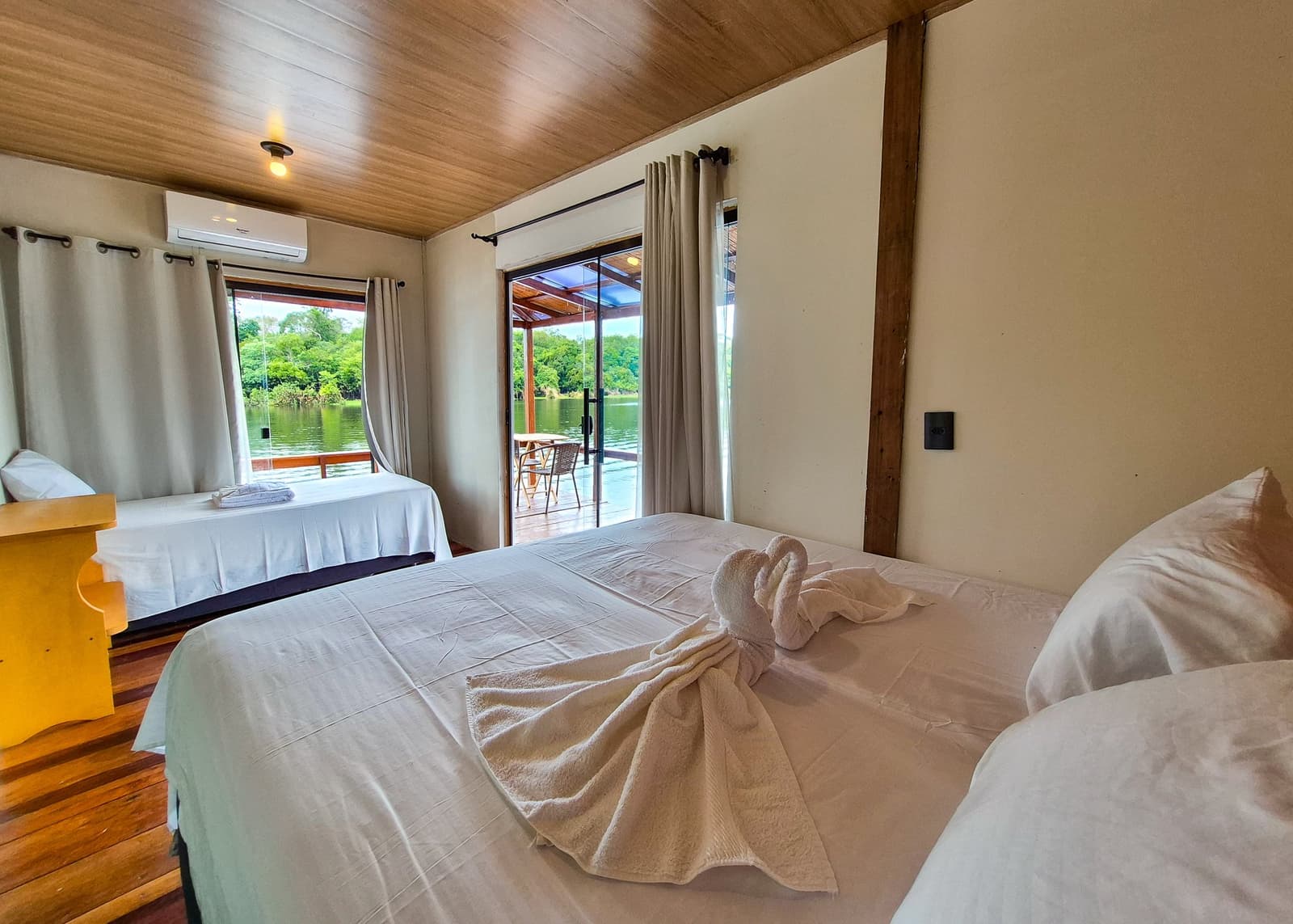 Fresh sheets, folded towels, and a river view waiting on the other side of the glass. Twin bedroom in an amazon jungle lodge with white linen beds, towel art on the double bed, wooden ceiling, air conditioning, and a sliding glass door opening onto a deck over a calm river flanked by dense green forest