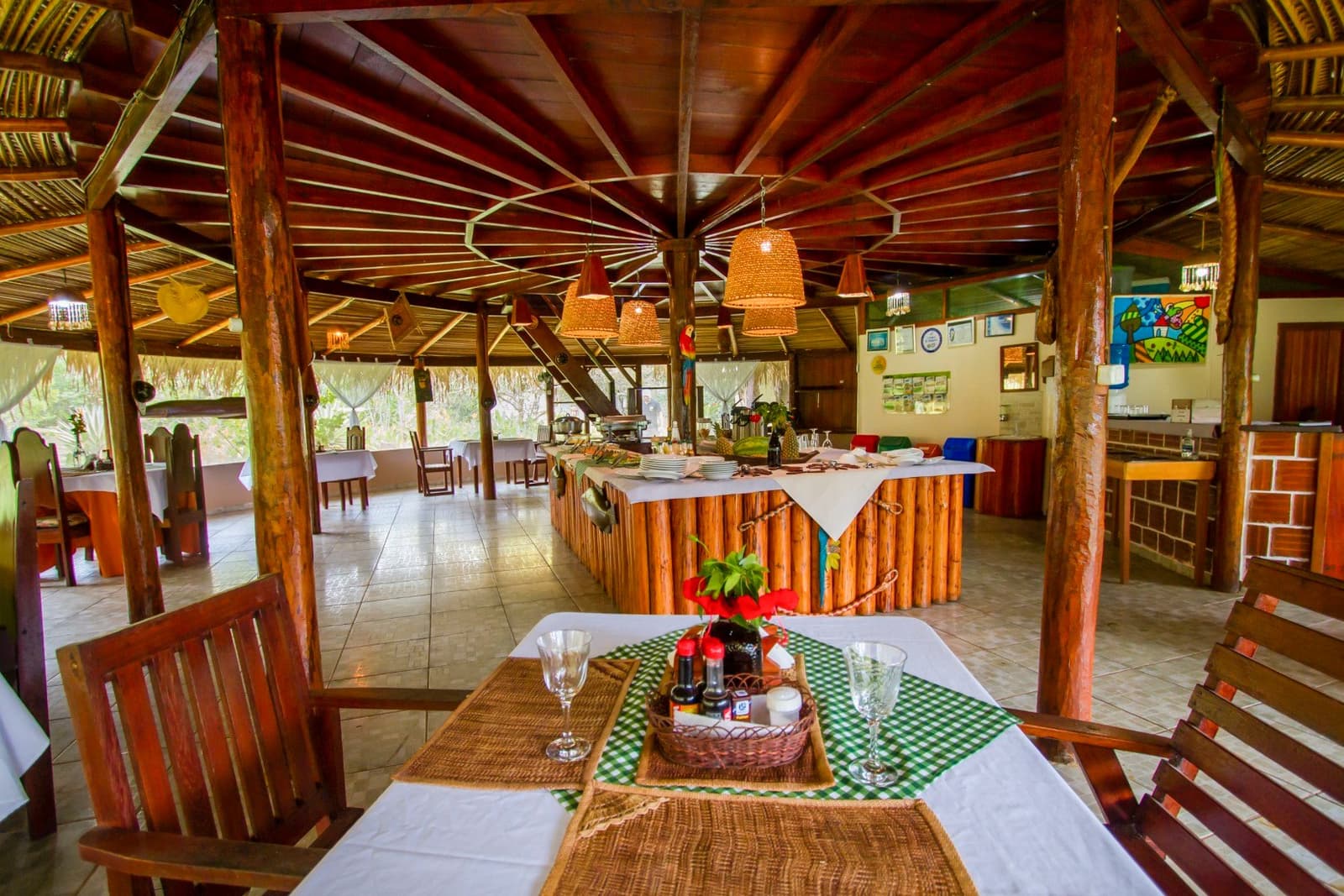 Dinner is served, and the wetlands are right outside the window. Open-air thatched dining room at a Pantanal lodge on an amazon and pantanal tour, with rustic log-trimmed buffet counter, wicker pendant lamps, white-clothed tables set with wine glasses and a wicker condiment basket, and jungle views through the open sides