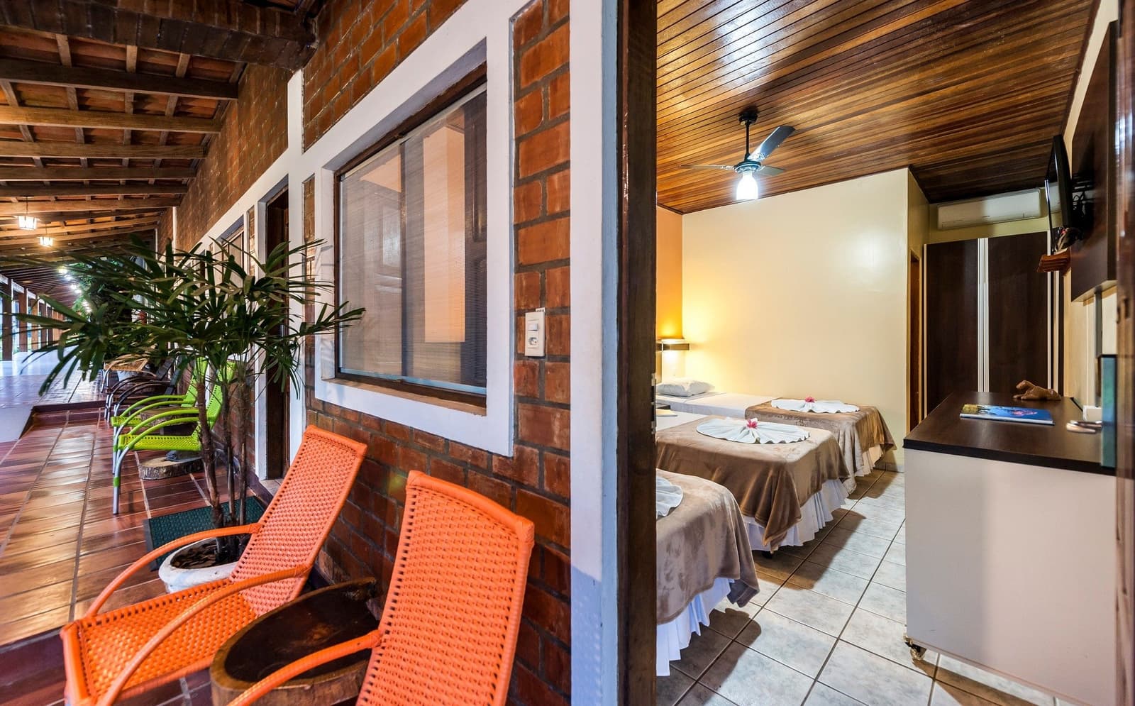 Good beds, a ceiling fan, and a chair outside. Honestly, that covers it. Open-air brick veranda with orange rattan chairs leading to a tidy twin room with wooden ceiling fan, warm bedside lighting, and towel-folded decorations on the beds, at a ranch-style pousada on an amazon and pantanal tour