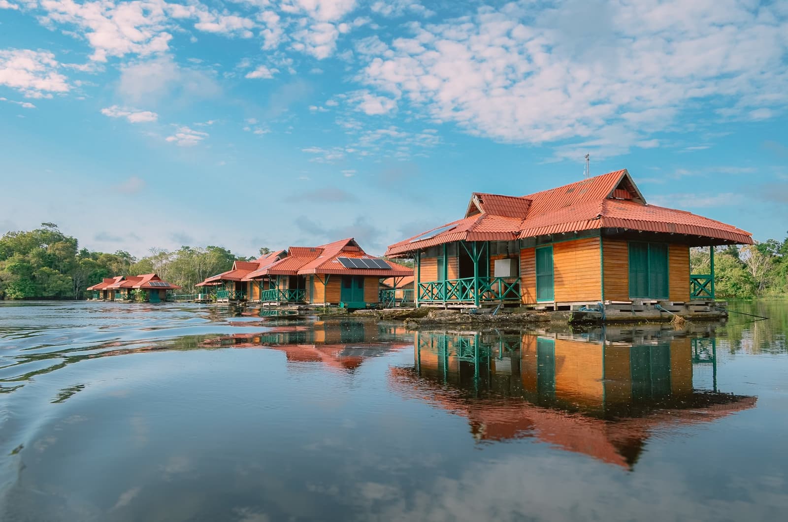 Your room for the next few nights, and it floats. A row of solar-powered wooden cabins with red roofs floating on a calm river during an amazon rainforest ecolodge trip in brazil, their warm orange walls reflected in the still dark water, dense green forest lining the far bank under a blue sky